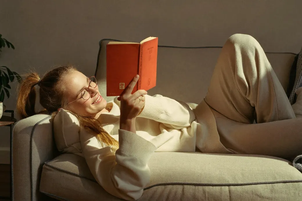 A woman stretched out on the couch reading and smiling, a slow Sunday morning in the sunlight